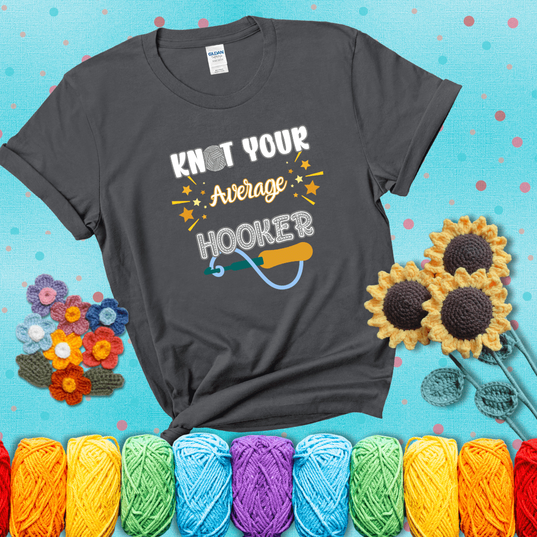 Bold lettering with a yarn hook graphic. Text reads: “I'm Not Your Average Hooker.” A cheeky crochet tee for confident crafters who own their skills, showing a dark grey tee with a teal sunflower background