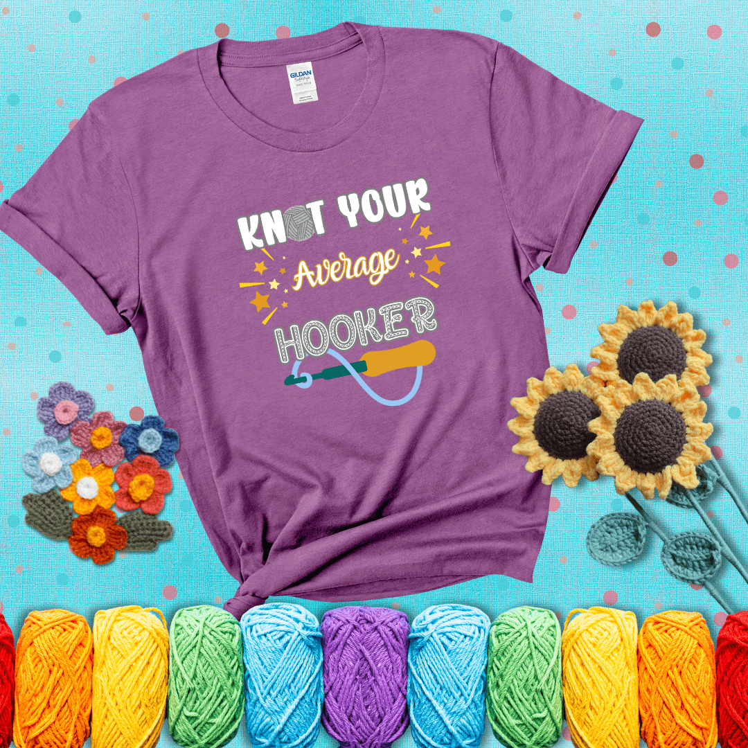 Bold lettering with a yarn hook graphic. Text reads: “I'm Not Your Average Hooker.” A cheeky crochet tee for confident crafters who own their skills, showing a light purple tee with a teal sunflower background