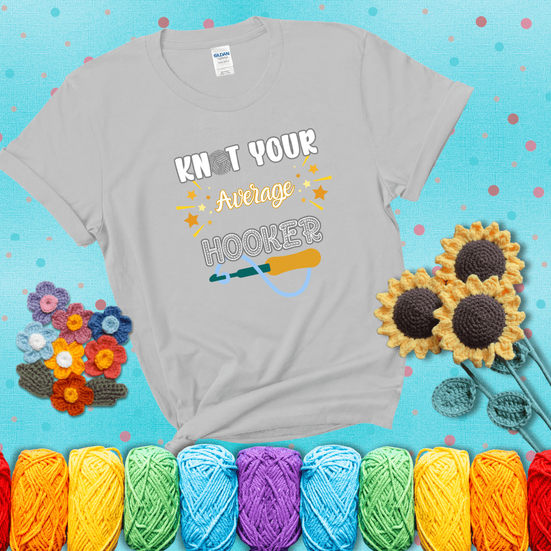 Bold lettering with a yarn hook graphic. Text reads: “I'm Not Your Average Hooker.” A cheeky crochet tee for confident crafters who own their skills, showing a light grey tee with a teal sunflower background