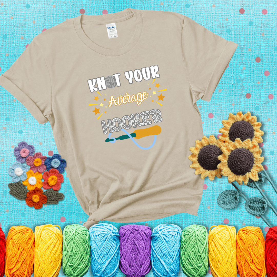 Bold lettering with a yarn hook graphic. Text reads: “I'm Not Your Average Hooker.” A cheeky crochet tee for confident crafters who own their skills, showing a beige tee with a teal sunflower background