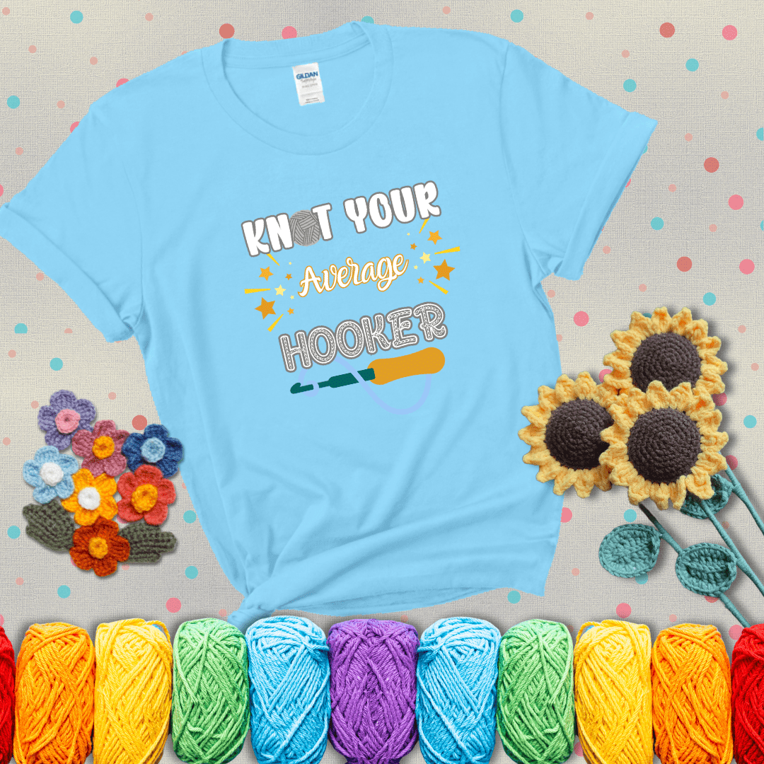 Bold lettering with a yarn hook graphic. Text reads: “I'm Not Your Average Hooker.” A cheeky crochet tee for confident crafters who own their skills, showing a light blue tee with grey sunflower background
