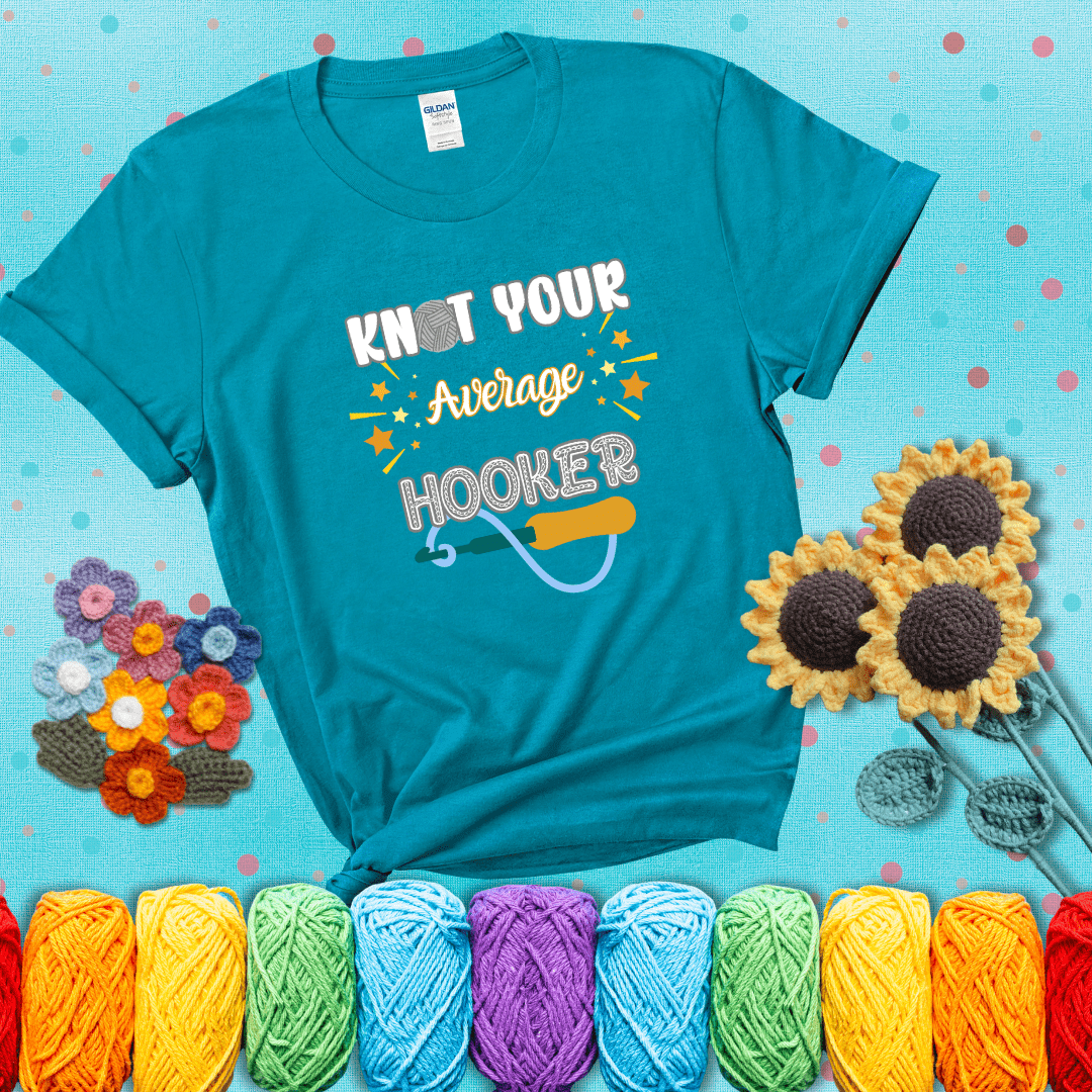 Bold lettering with a yarn hook graphic. Text reads: “I'm Not Your Average Hooker.” A cheeky crochet tee for confident crafters who own their skills, showing a blue-green tee with a teal sunflower background