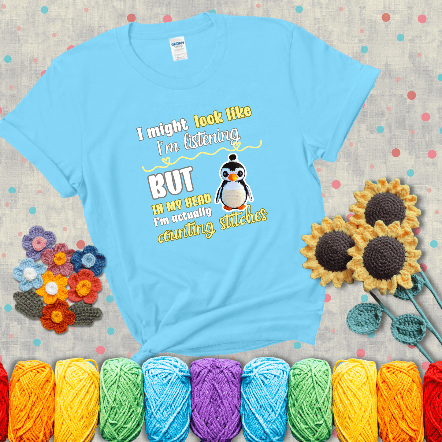 Crochet penguin next to the text “I might look like I’m listening but in my head I’m actually counting stitches” showing a light blue tee with grey sunflower background