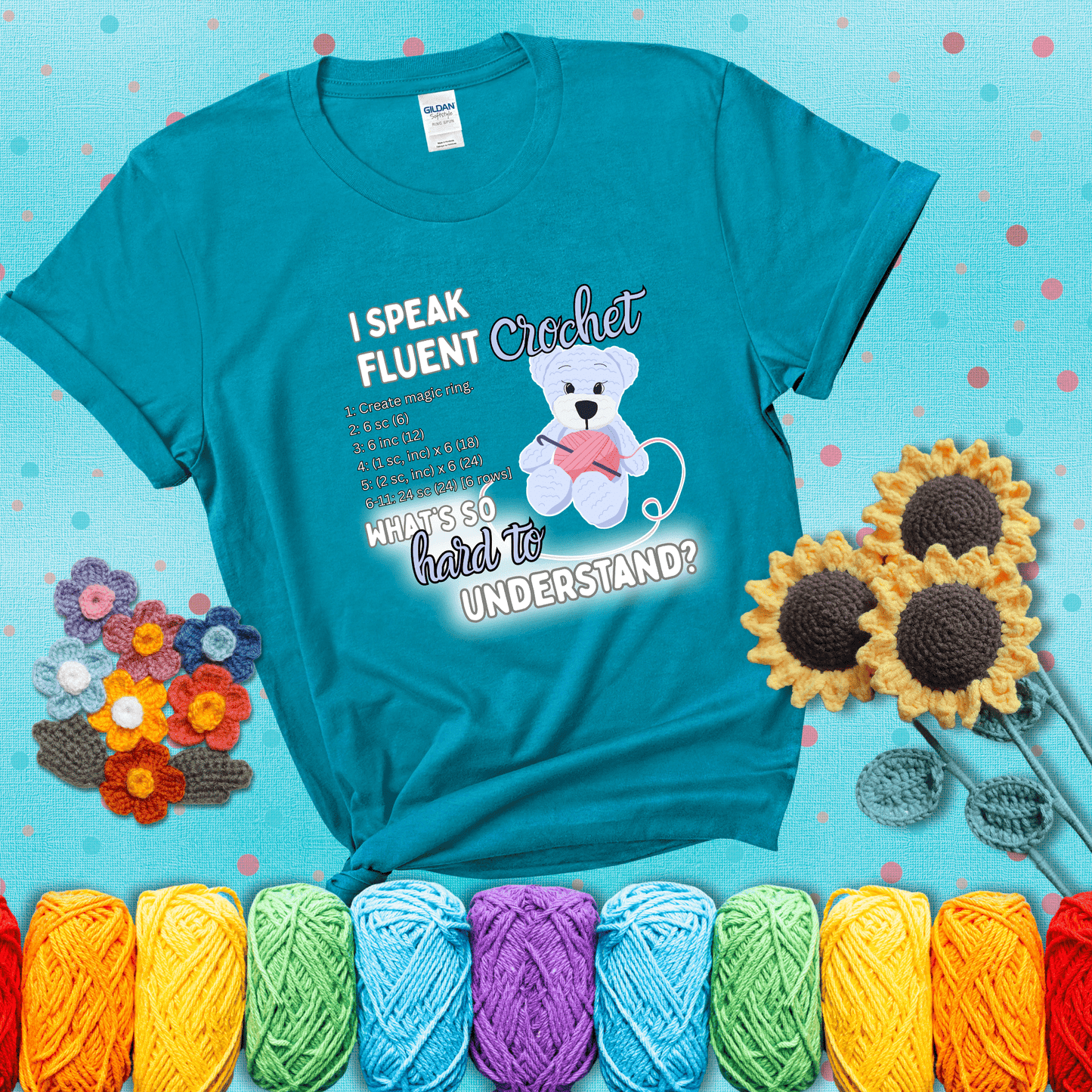 A cute crochet bear holding a yarn ball, surrounded by a short crochet pattern. The phrase reads “I Speak Fluent Crochet – What’s so hard to understand?” showing a blue-green tee with a teal sunflower background