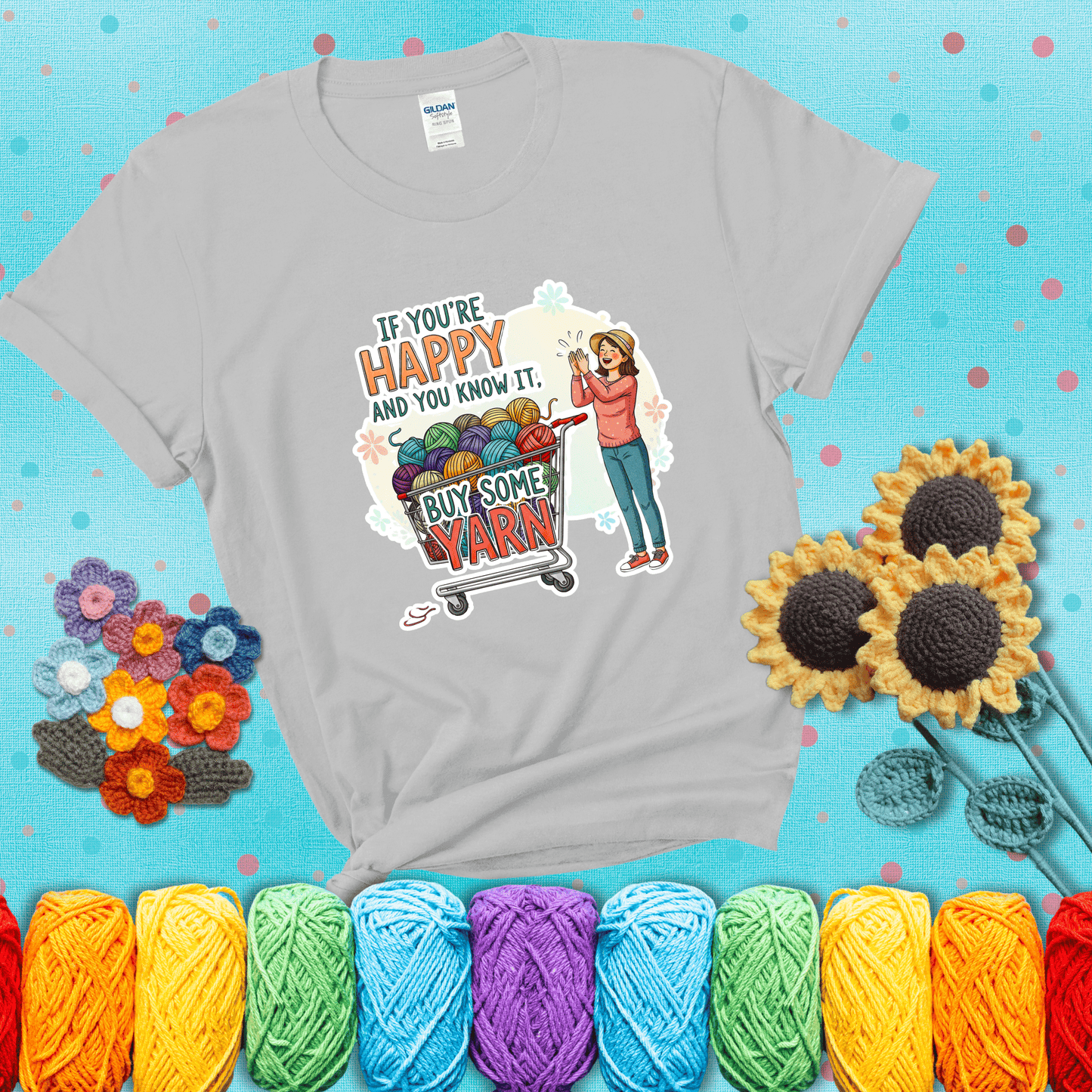 Woman clapping beside a shopping cart overflowing with colorful yarn balls, styled with cheerful pastel accents and bold playful lettering showing a light grey tee with a teal sunflower background