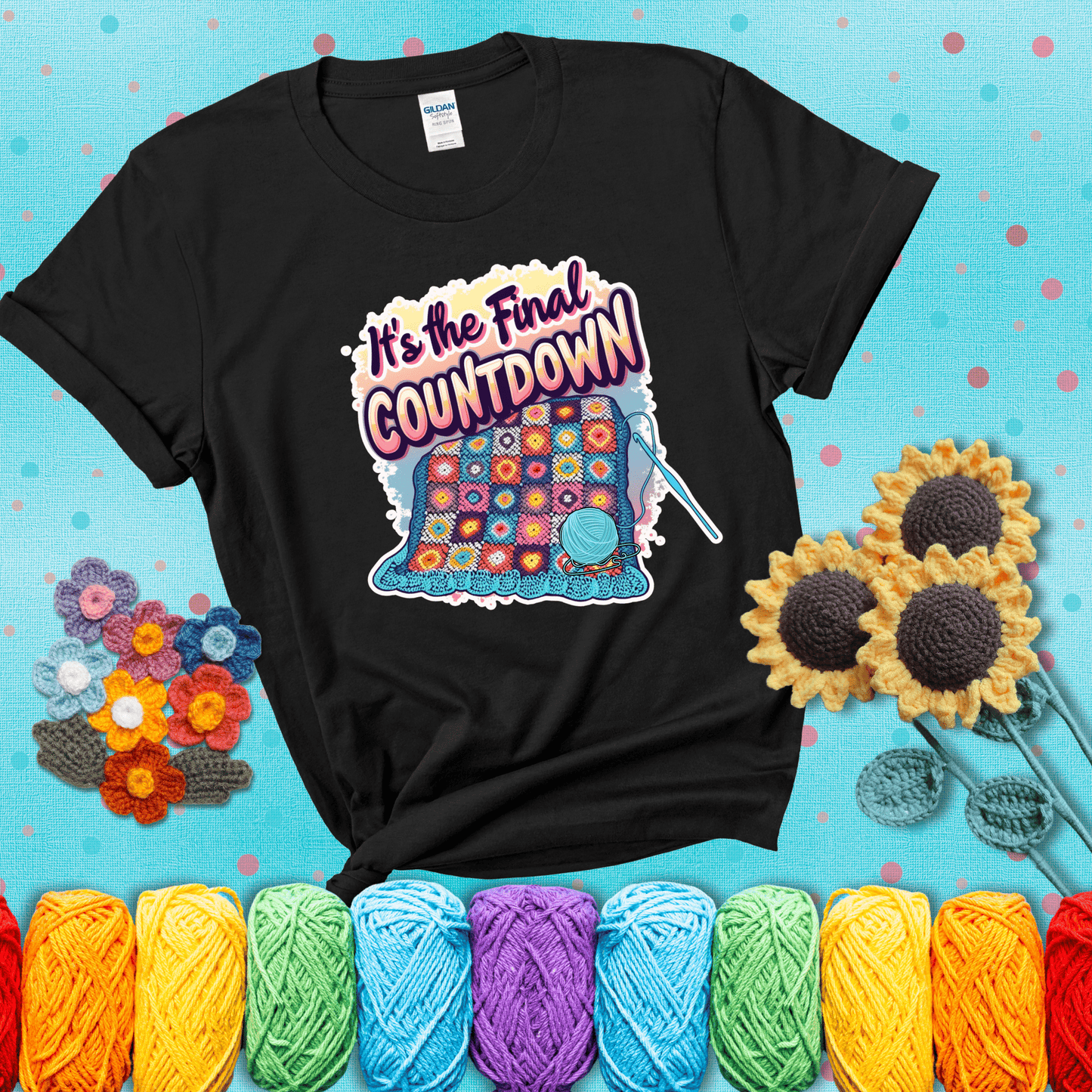 Colorful crochet blanket in progress with text "It’s the Final Countdown" above it in bold, multicolor lettering. A blue ball of yarn and hook are placed at the bottom, showing a black tee with a teal sunflower background