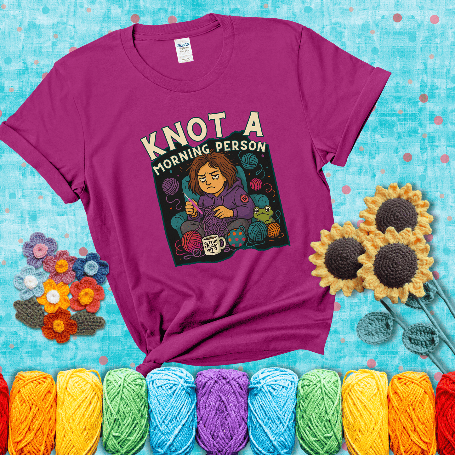 Sleepy crocheter in a purple hoodie sits in a chair with tangled yarn, coffee mug, and a frog sidekick, surrounded by yarn balls showing a magenta tee with a teal sunflower background