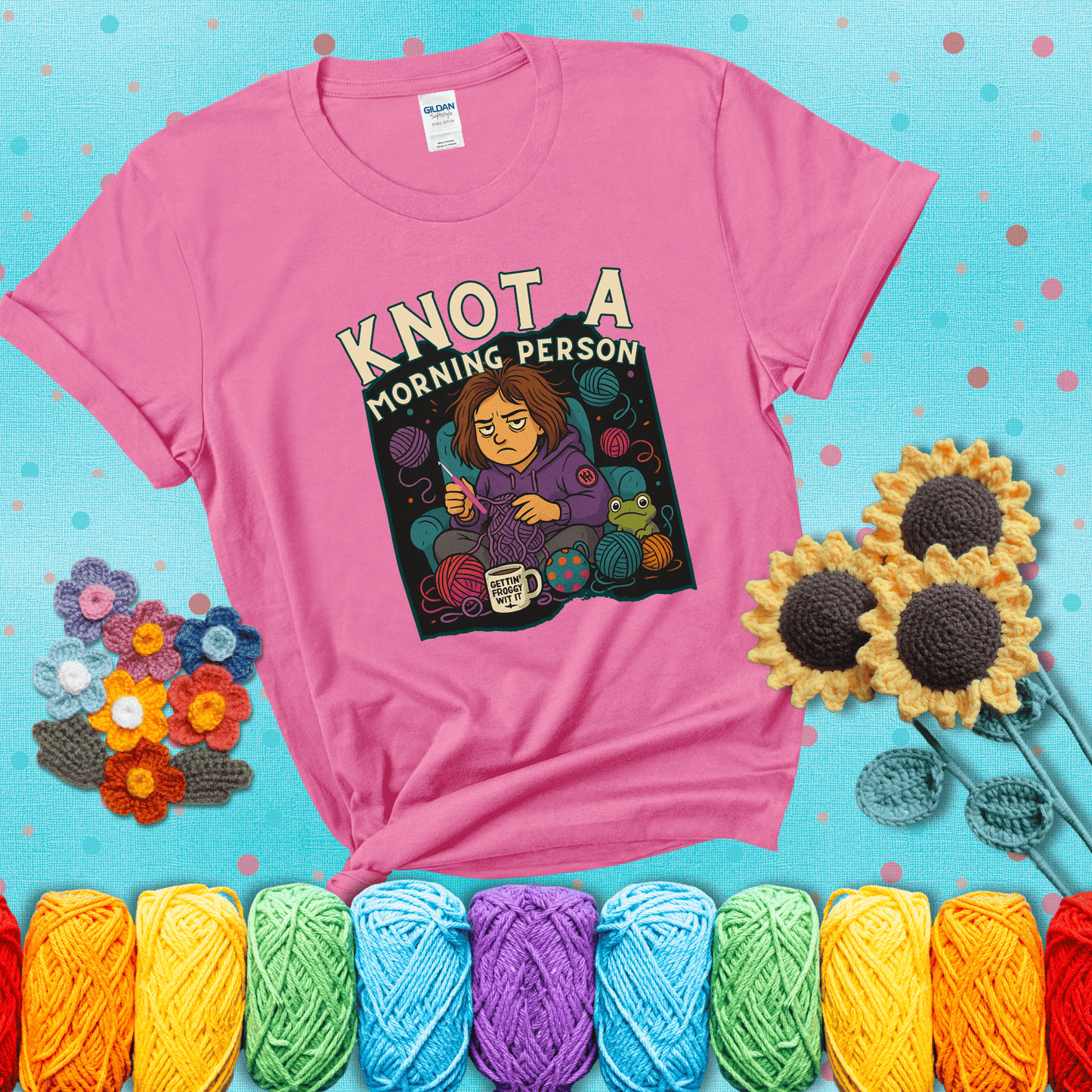 Sleepy crocheter in a purple hoodie sits in a chair with tangled yarn, coffee mug, and a frog sidekick, surrounded by yarn balls showing a pink tee with a teal sunflower background