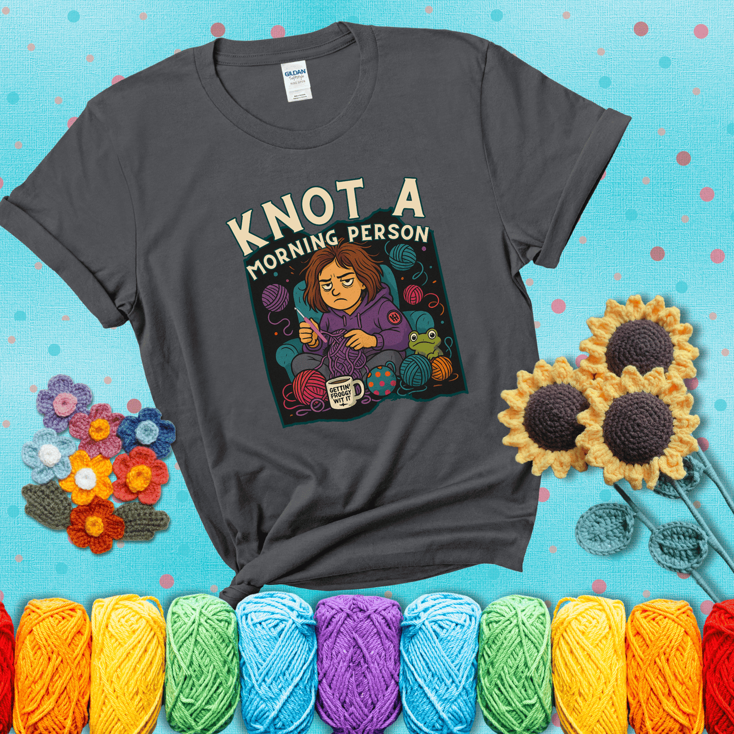 Sleepy crocheter in a purple hoodie sits in a chair with tangled yarn, coffee mug, and a frog sidekick, surrounded by yarn balls showing a dark grey tee with a teal sunflower background