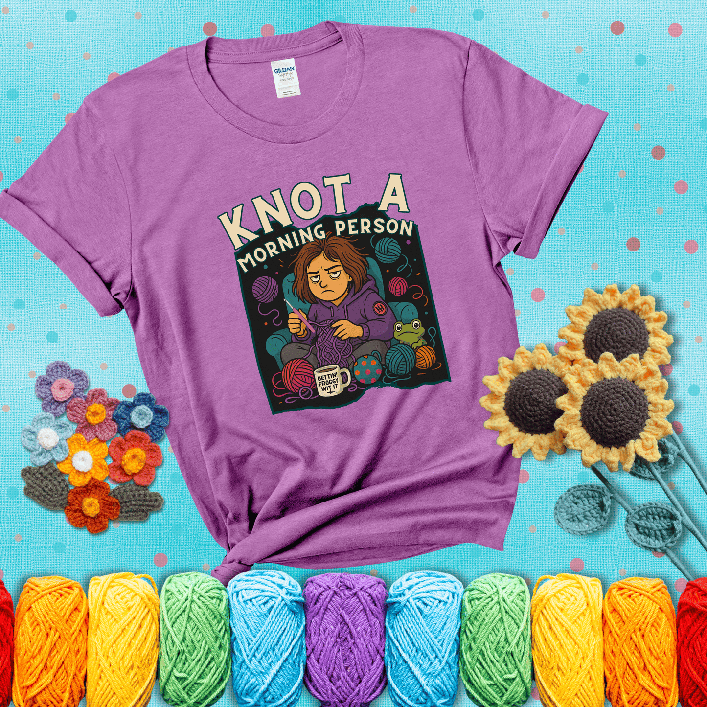Sleepy crocheter in a purple hoodie sits in a chair with tangled yarn, coffee mug, and a frog sidekick, surrounded by yarn balls showing a light purple tee with a teal sunflower background