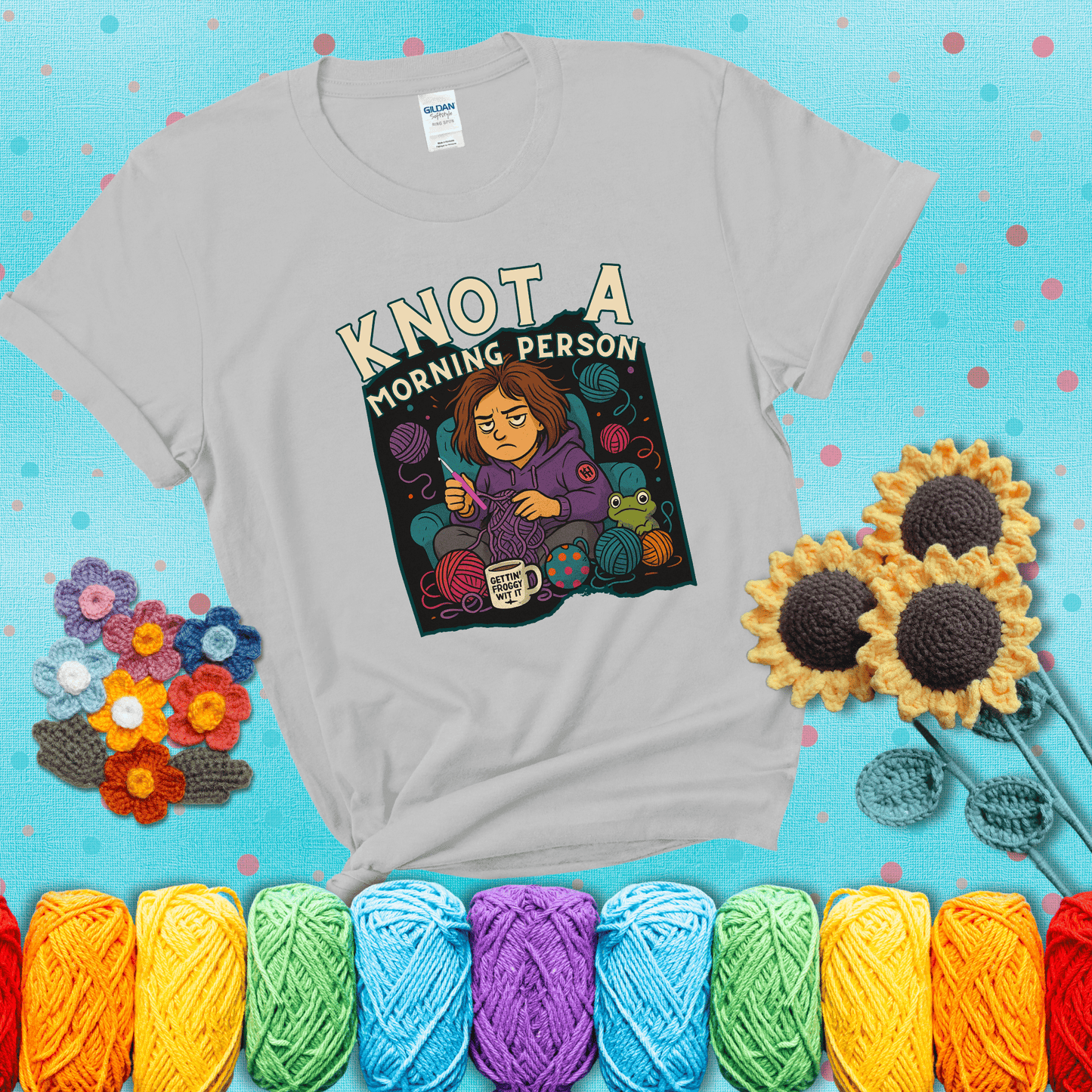 Sleepy crocheter in a purple hoodie sits in a chair with tangled yarn, coffee mug, and a frog sidekick, surrounded by yarn balls showing a light grey tee with a teal sunflower background