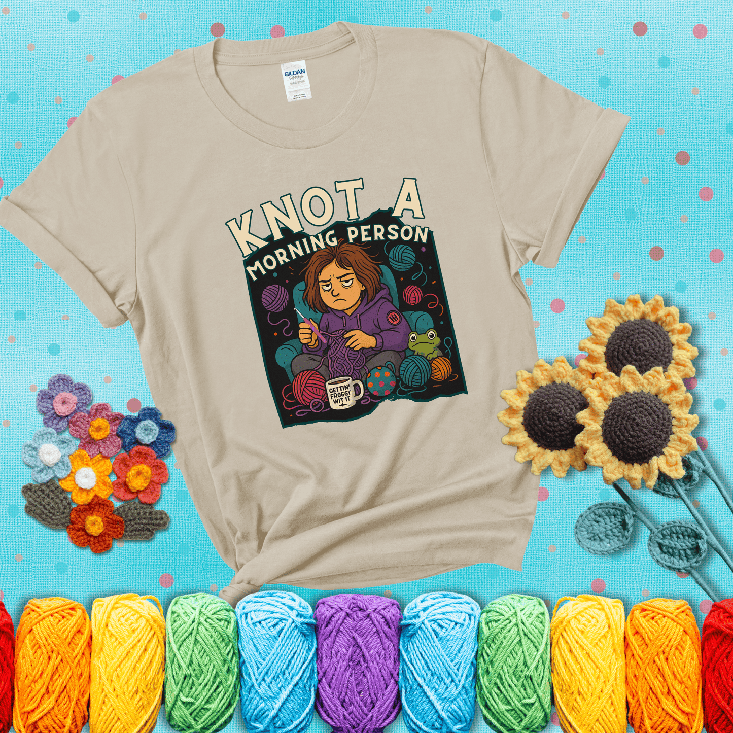 Sleepy crocheter in a purple hoodie sits in a chair with tangled yarn, coffee mug, and a frog sidekick, surrounded by yarn balls showing a beige tee with a teal sunflower background
