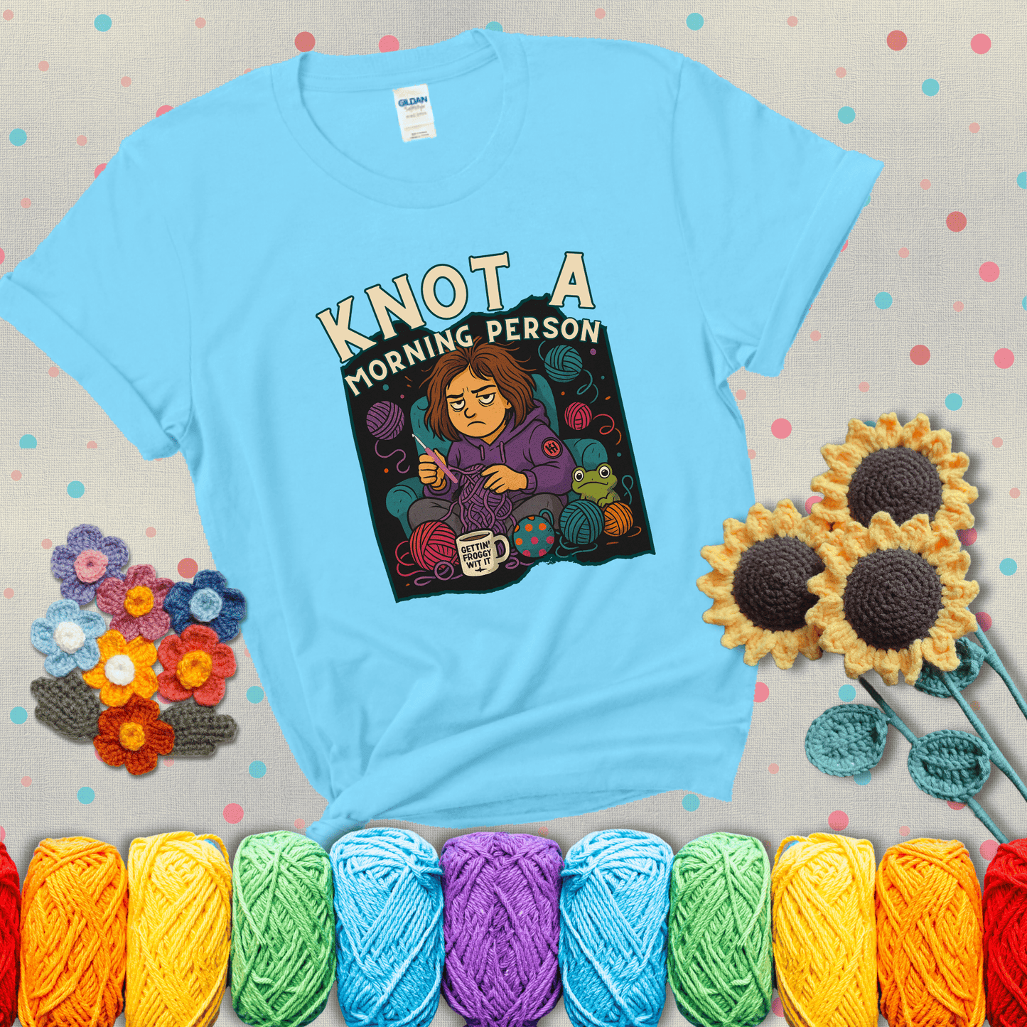 Sleepy crocheter in a purple hoodie sits in a chair with tangled yarn, coffee mug, and a frog sidekick, surrounded by yarn balls showing a light blue tee with grey sunflower background