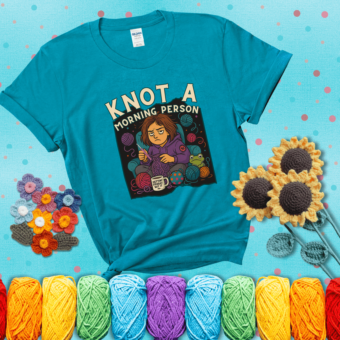 Sleepy crocheter in a purple hoodie sits in a chair with tangled yarn, coffee mug, and a frog sidekick, surrounded by yarn balls showing a blue-green tee with a teal sunflower background