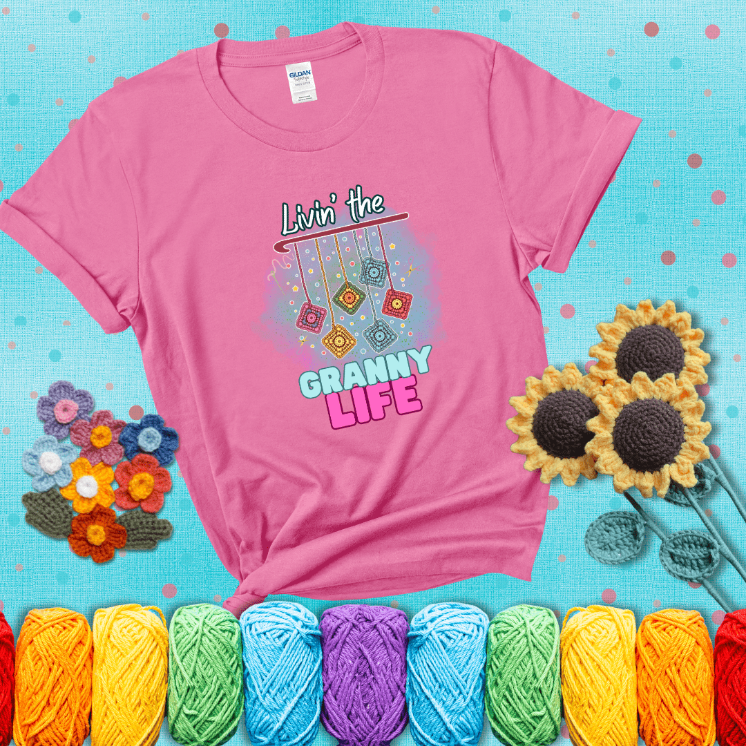 Colorful crochet granny squares hang from a hook with text “Livin’ the Granny Life” in bold pink and blue font, a fun nod to classic crochet showing a pink tee with a teal sunflower background