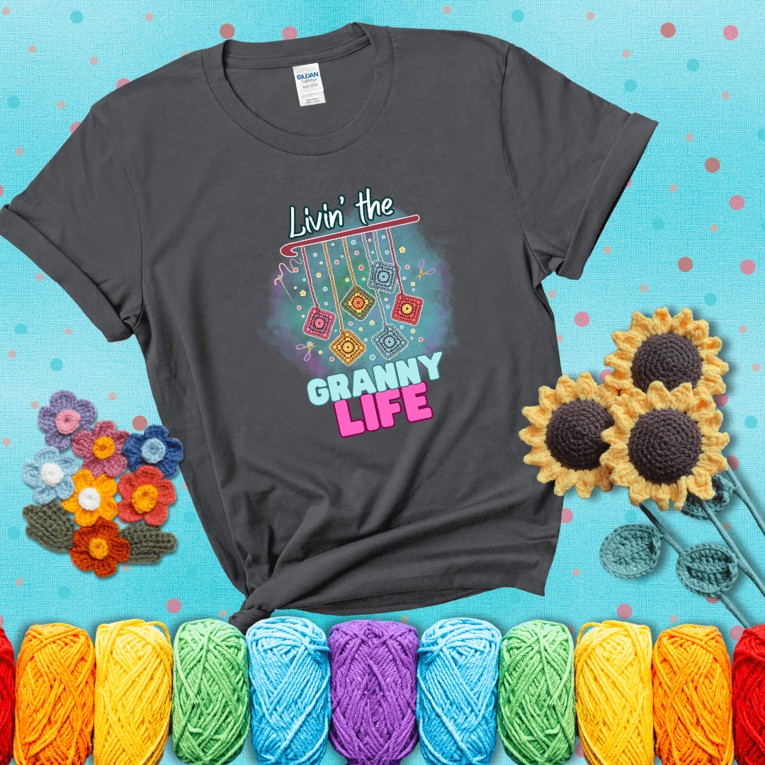 Colorful crochet granny squares hang from a hook with text “Livin’ the Granny Life” in bold pink and blue font, a fun nod to classic crochet showing a dark grey tee with a teal sunflower background
