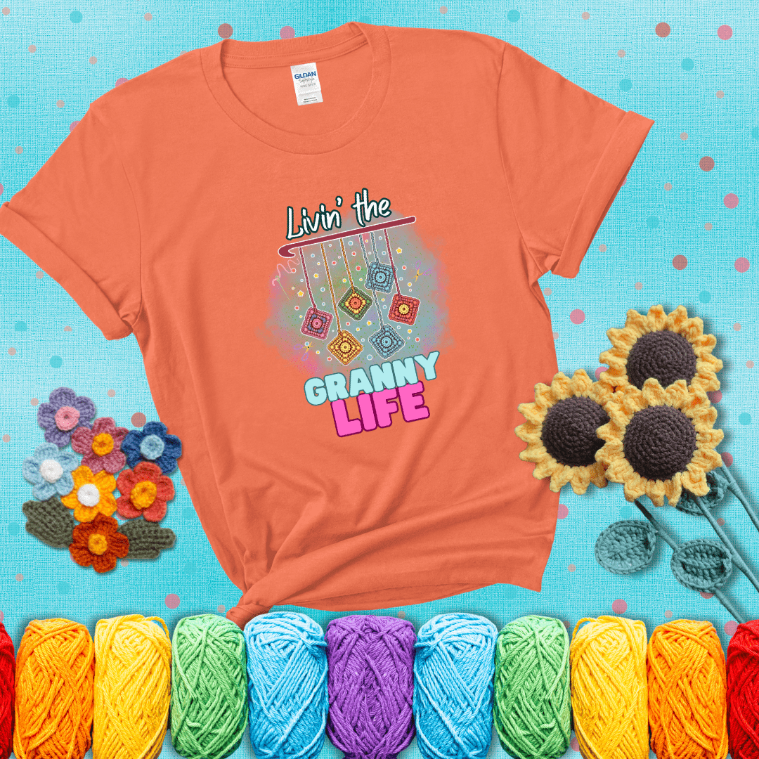 Colorful crochet granny squares hang from a hook with text “Livin’ the Granny Life” in bold pink and blue font, a fun nod to classic crochet showing a light orange tee with a teal sunflower background