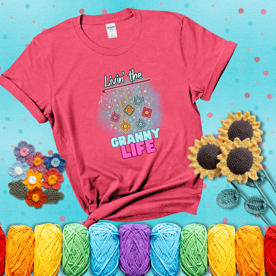 Colorful crochet granny squares hang from a hook with text “Livin’ the Granny Life” in bold pink and blue font, a fun nod to classic crochet showing a light red tee with a teal sunflower background