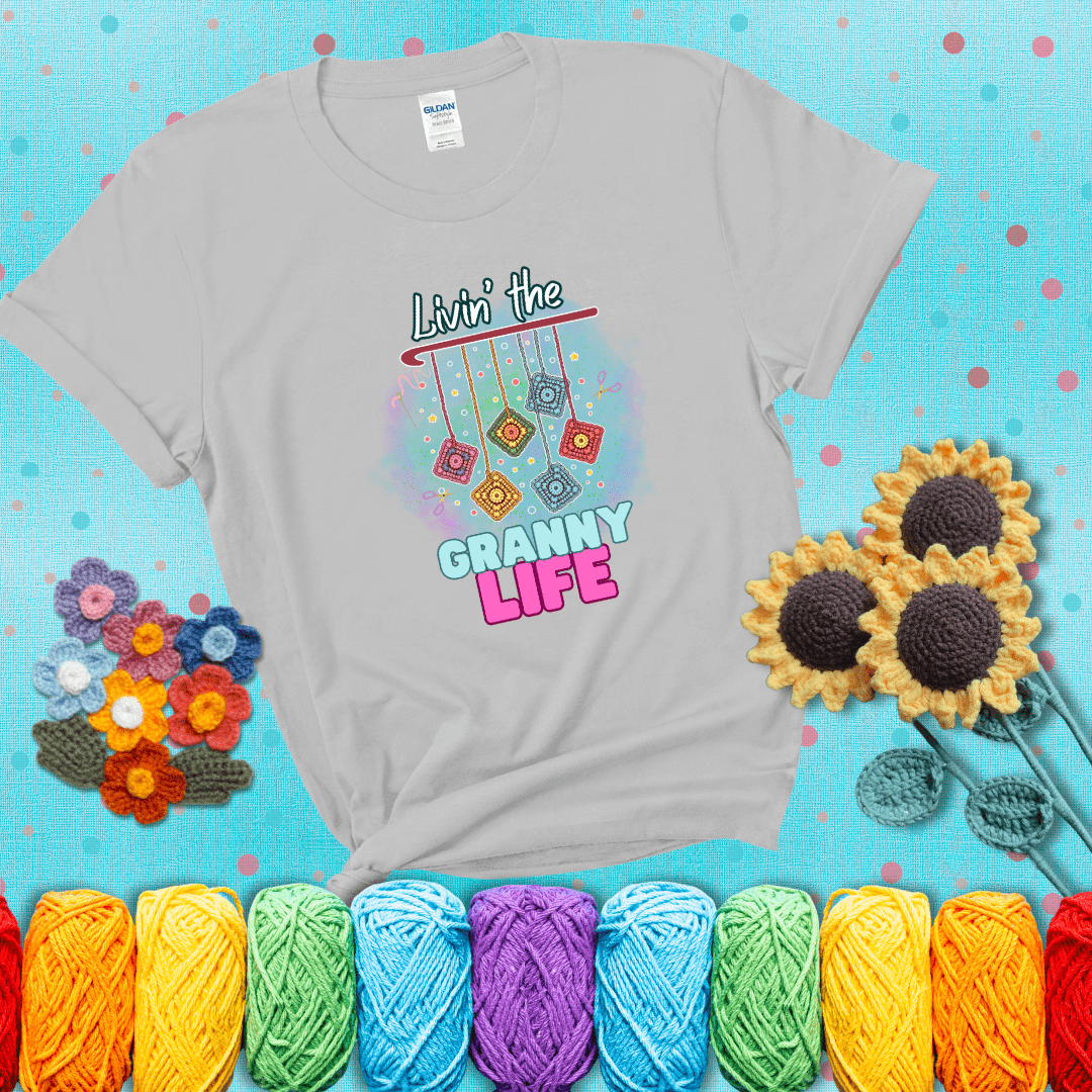 Colorful crochet granny squares hang from a hook with text “Livin’ the Granny Life” in bold pink and blue font, a fun nod to classic crochet showing a light grey tee with a teal sunflower background