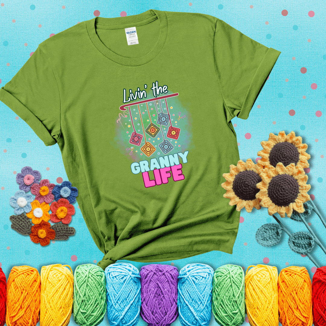 Colorful crochet granny squares hang from a hook with text “Livin’ the Granny Life” in bold pink and blue font, a fun nod to classic crochet showing a bright green tee with a teal sunflower background