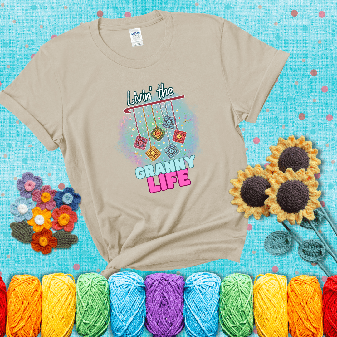 Colorful crochet granny squares hang from a hook with text “Livin’ the Granny Life” in bold pink and blue font, a fun nod to classic crochet showing a beige tee with a teal sunflower background