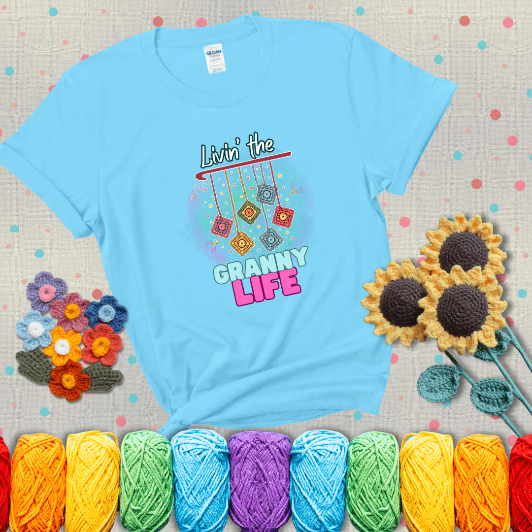 Colorful crochet granny squares hang from a hook with text “Livin’ the Granny Life” in bold pink and blue font, a fun nod to classic crochet showing a light blue tee with grey sunflower background