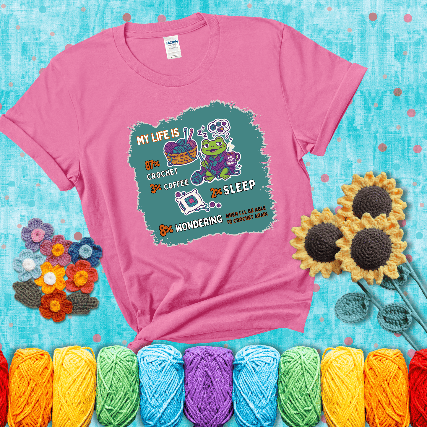 A relaxed frog crocheting while sipping coffee, surrounded by yarn and dreaming of more yarn. Includes whimsical text dividing life into percentages: 87% crochet, 3% coffee, 2% sleep, 8% wondering when to crochet again showing a pink tee with a teal sunflower background