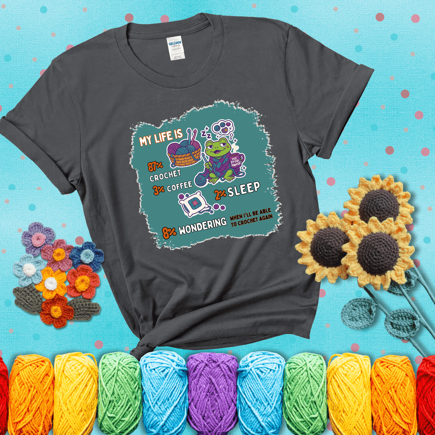 A relaxed frog crocheting while sipping coffee, surrounded by yarn and dreaming of more yarn. Includes whimsical text dividing life into percentages: 87% crochet, 3% coffee, 2% sleep, 8% wondering when to crochet again showing a dark grey tee with a teal sunflower background