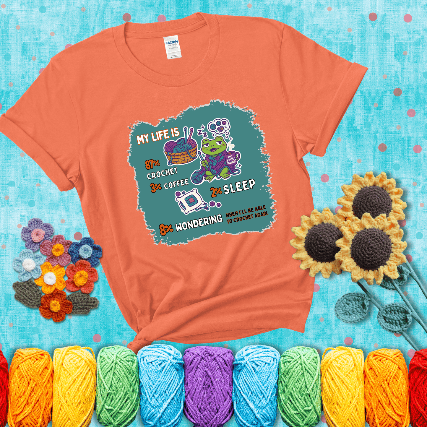 A relaxed frog crocheting while sipping coffee, surrounded by yarn and dreaming of more yarn. Includes whimsical text dividing life into percentages: 87% crochet, 3% coffee, 2% sleep, 8% wondering when to crochet again showing a light orange tee with a teal sunflower background