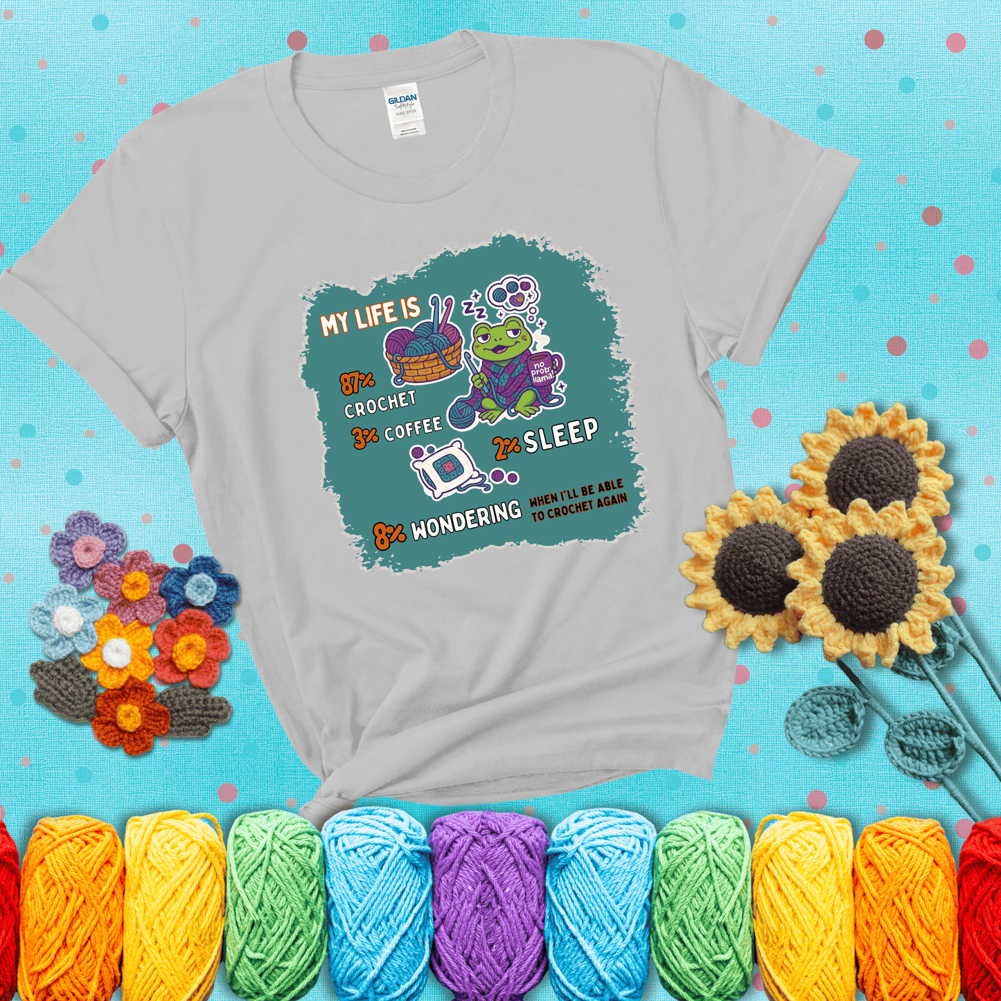 A relaxed frog crocheting while sipping coffee, surrounded by yarn and dreaming of more yarn. Includes whimsical text dividing life into percentages: 87% crochet, 3% coffee, 2% sleep, 8% wondering when to crochet again showing a light grey tee with a teal sunflower background