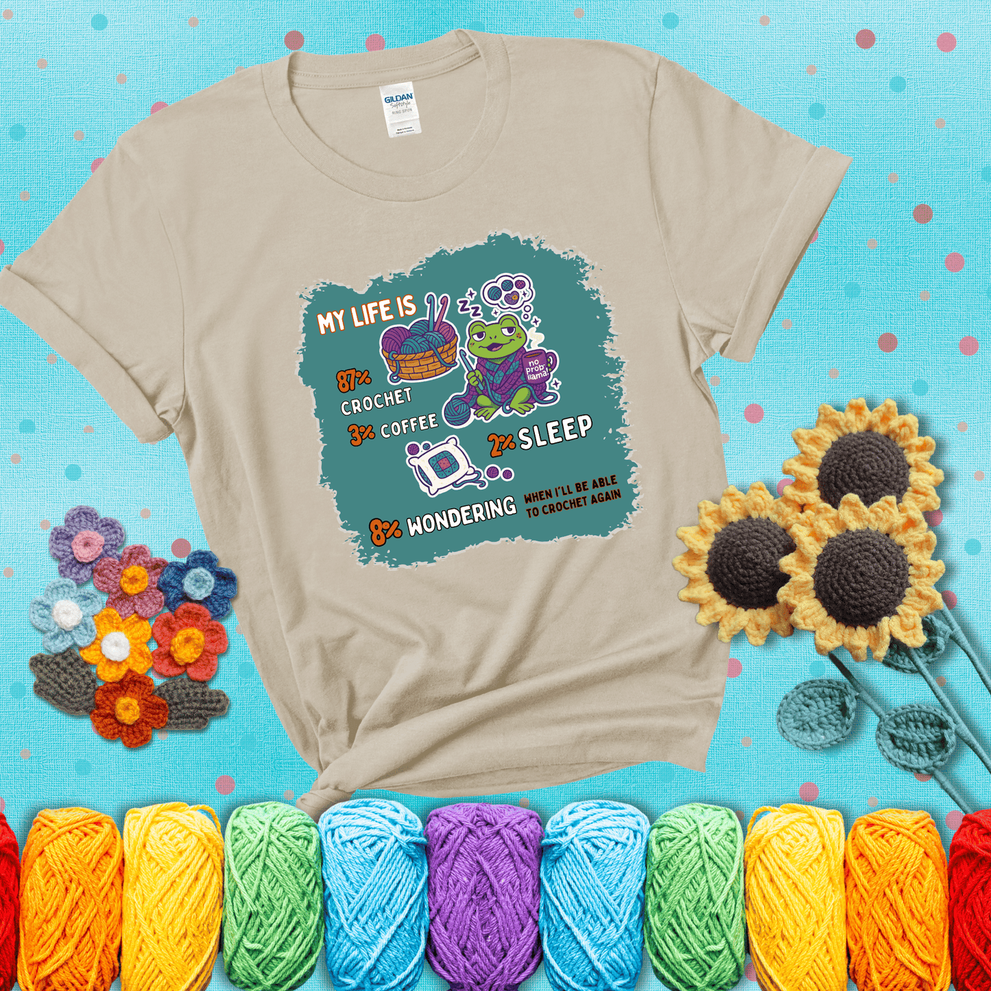 A relaxed frog crocheting while sipping coffee, surrounded by yarn and dreaming of more yarn. Includes whimsical text dividing life into percentages: 87% crochet, 3% coffee, 2% sleep, 8% wondering when to crochet again showing a beige tee with a teal sunflower background
