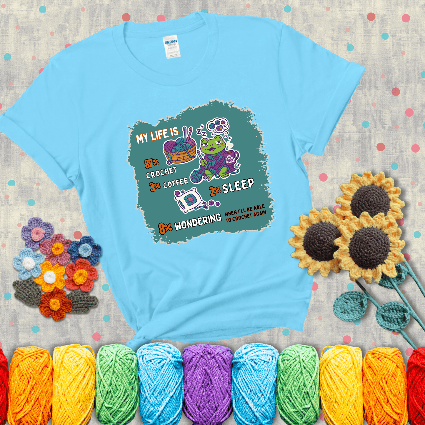 A relaxed frog crocheting while sipping coffee, surrounded by yarn and dreaming of more yarn. Includes whimsical text dividing life into percentages: 87% crochet, 3% coffee, 2% sleep, 8% wondering when to crochet again showing a light blue tee with grey sunflower background
