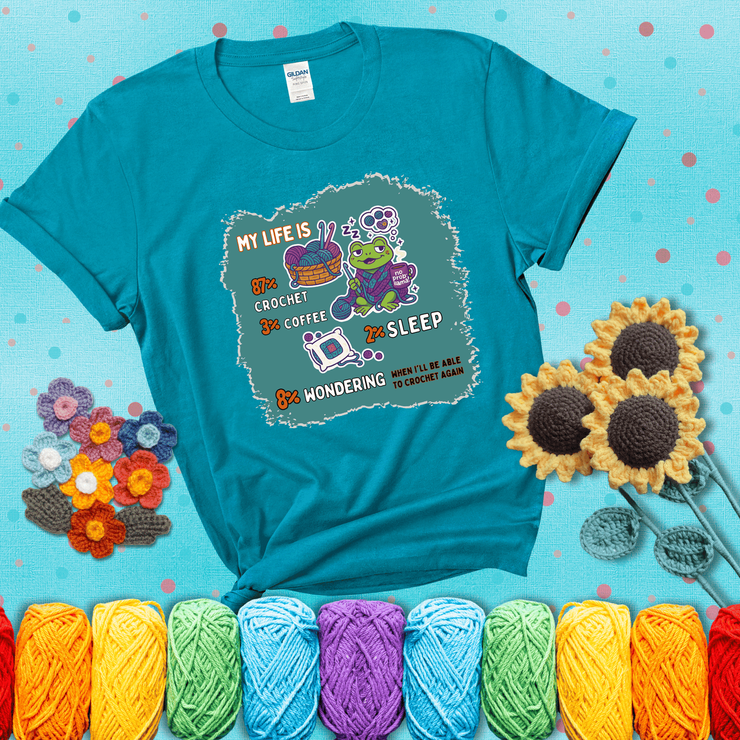 A relaxed frog crocheting while sipping coffee, surrounded by yarn and dreaming of more yarn. Includes whimsical text dividing life into percentages: 87% crochet, 3% coffee, 2% sleep, 8% wondering when to crochet again showing a blue-green tee with a teal sunflower background