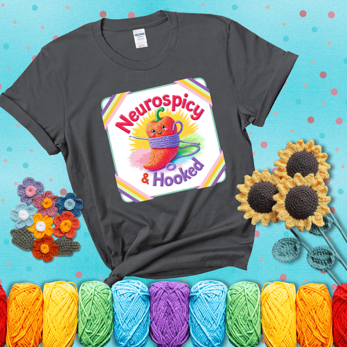 A cheerful red pepper wrapped in purple yarn with a crochet hook, framed by pastel rainbow corners and the words "Neurospicy & Hooked" showing a dark grey tee with a teal sunflower background