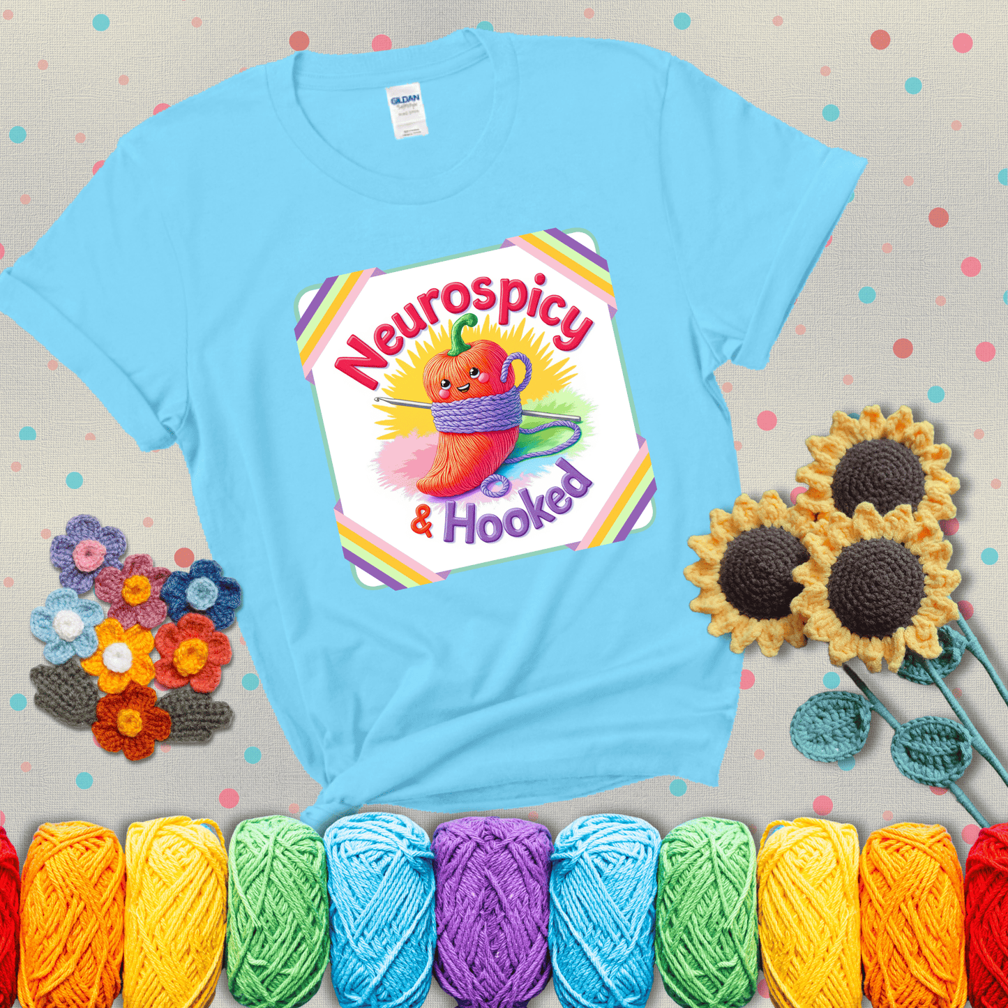 A cheerful red pepper wrapped in purple yarn with a crochet hook, framed by pastel rainbow corners and the words "Neurospicy & Hooked" showing a light blue tee with grey sunflower background