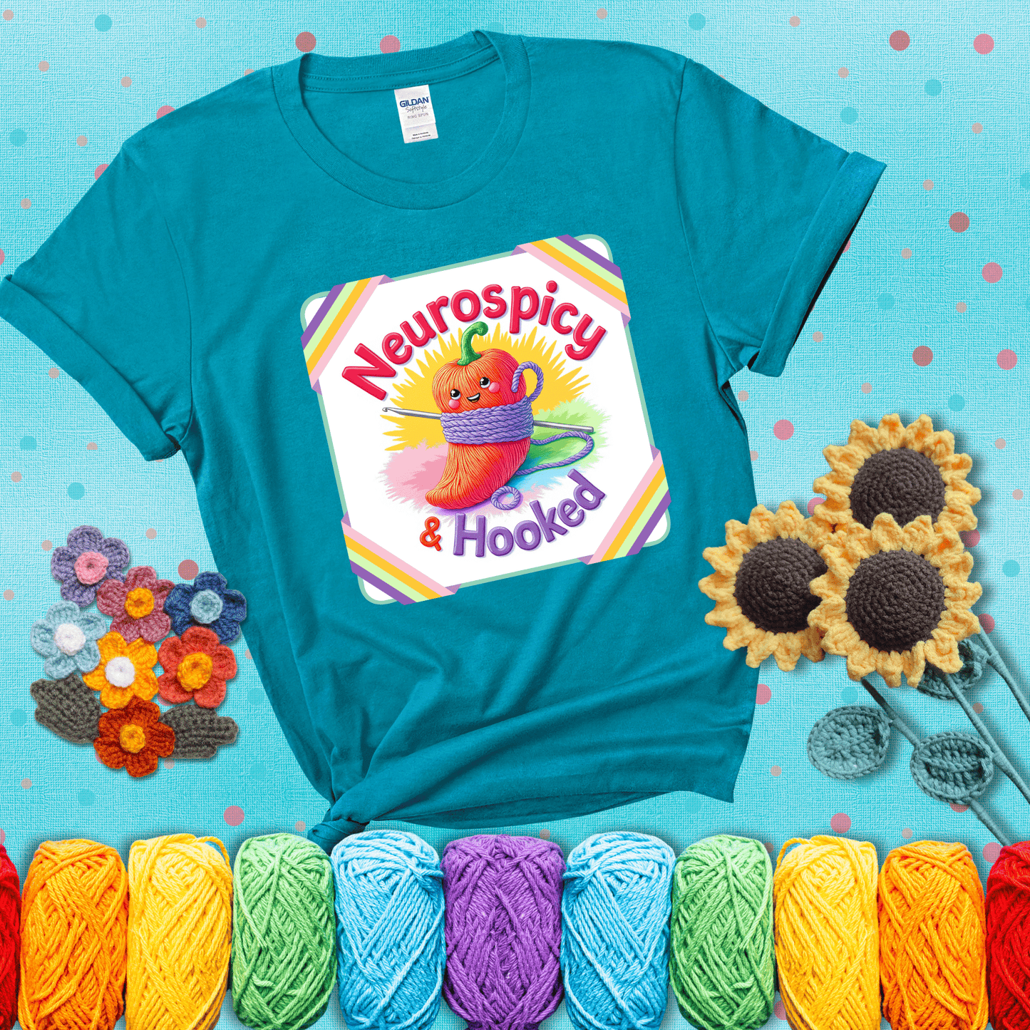A cheerful red pepper wrapped in purple yarn with a crochet hook, framed by pastel rainbow corners and the words "Neurospicy & Hooked" showing a blue-green tee with a teal sunflower background