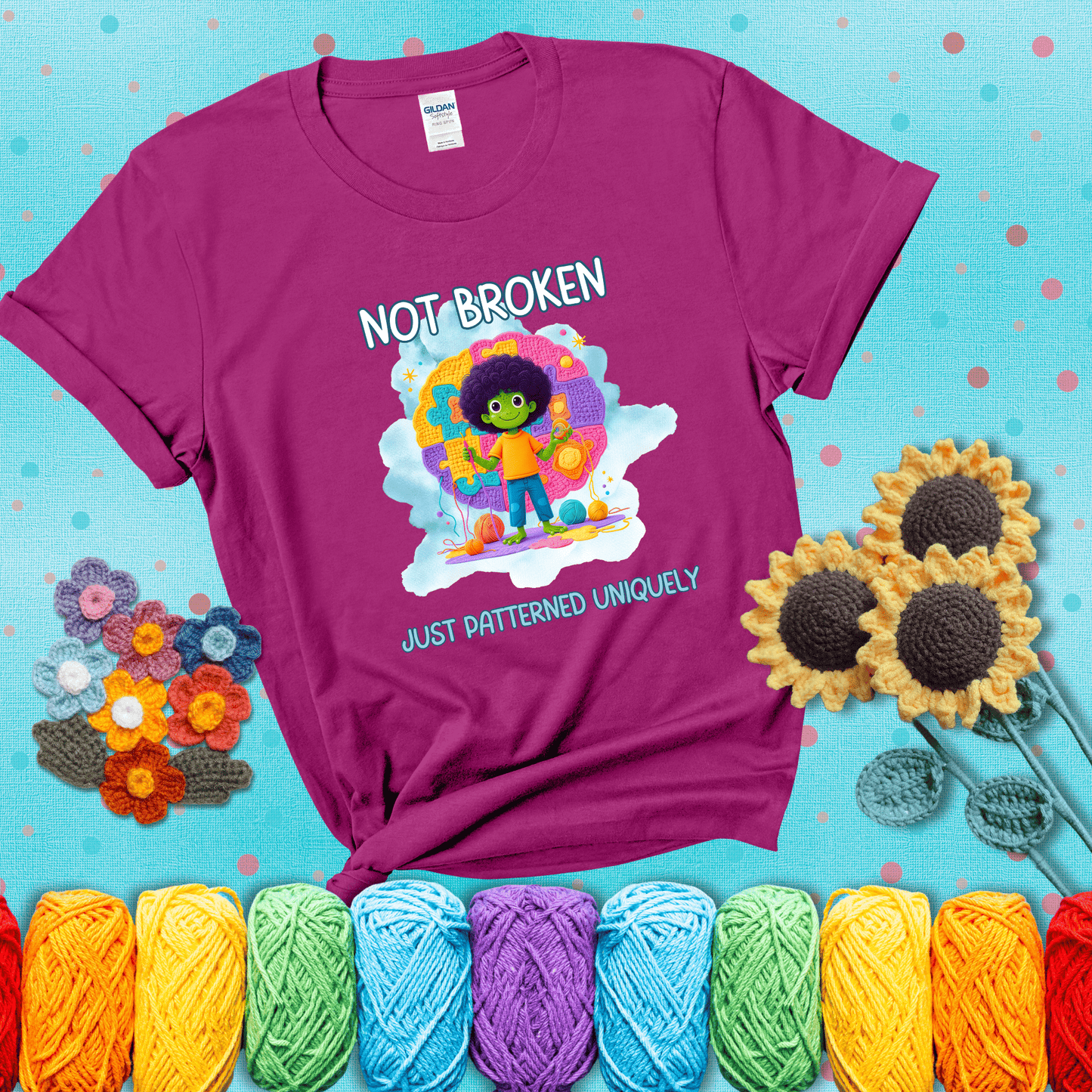 A smiling green character stands on a colorful crochet mat shaped like puzzle pieces, holding yarn and a hook, with the words “Not broken, just patterned uniquely” showing a magenta tee with a teal sunflower background