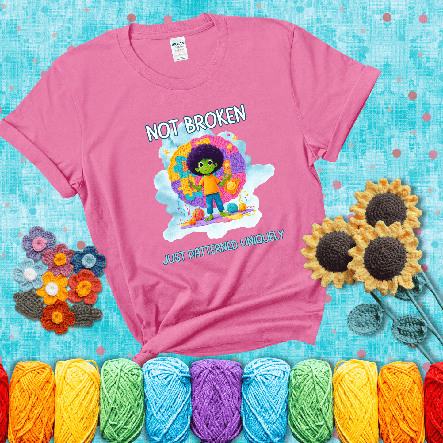 A smiling green character stands on a colorful crochet mat shaped like puzzle pieces, holding yarn and a hook, with the words “Not broken, just patterned uniquely” showing a pink tee with a teal sunflower background