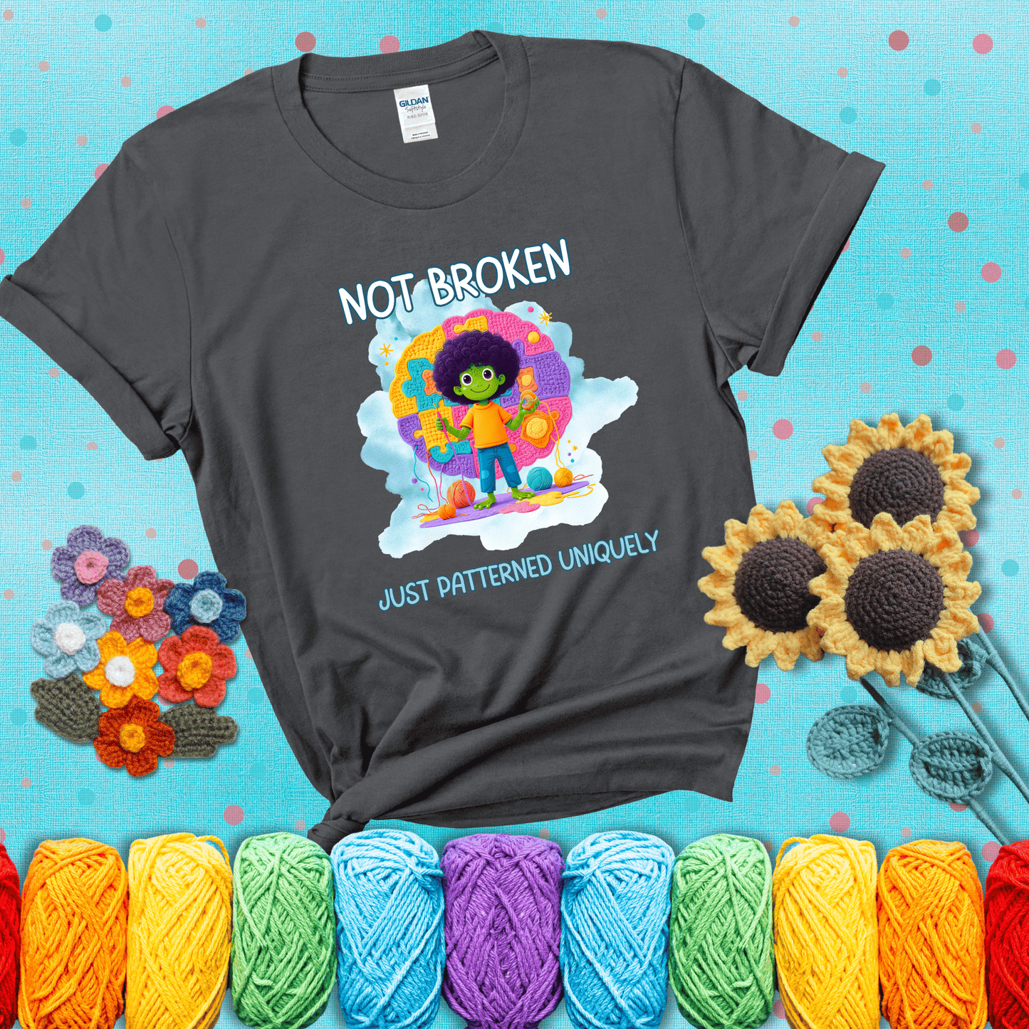 A smiling green character stands on a colorful crochet mat shaped like puzzle pieces, holding yarn and a hook, with the words “Not broken, just patterned uniquely” showing a dark grey tee with a teal sunflower background