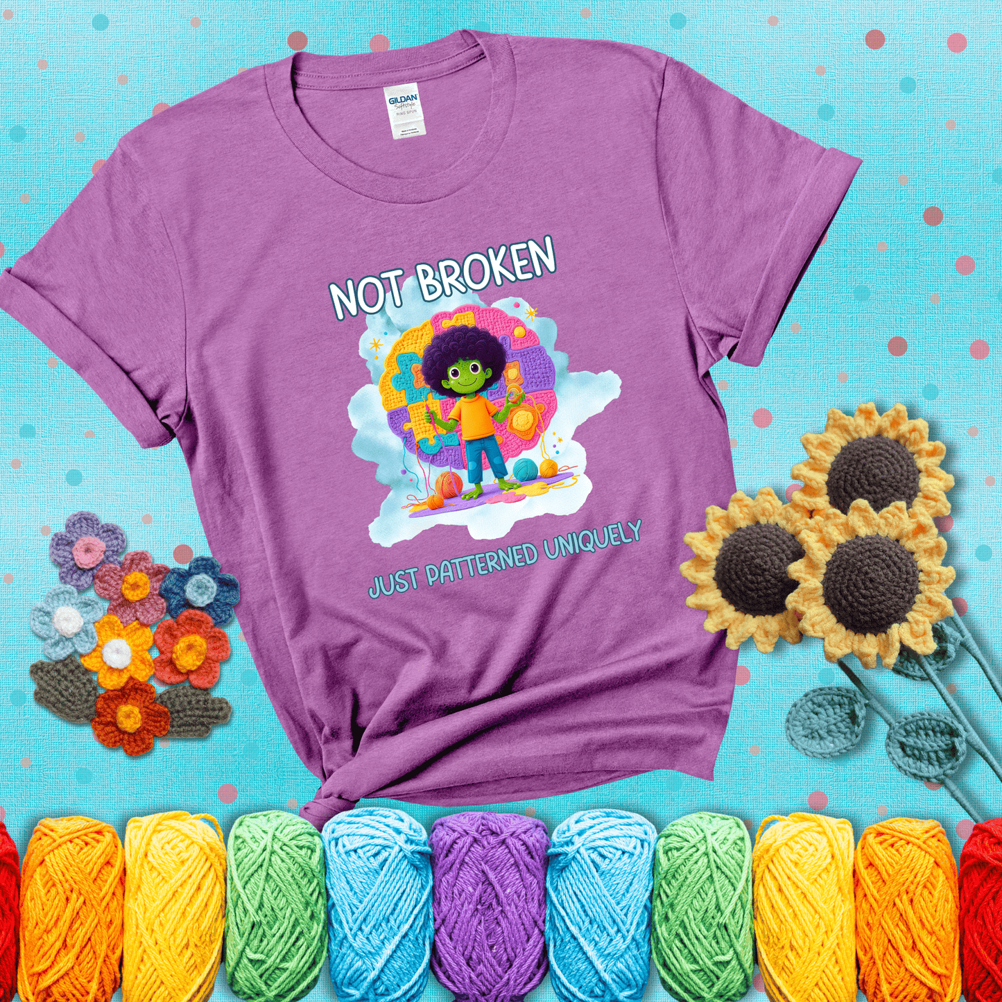 A smiling green character stands on a colorful crochet mat shaped like puzzle pieces, holding yarn and a hook, with the words “Not broken, just patterned uniquely” showing a light purple tee with a teal sunflower background