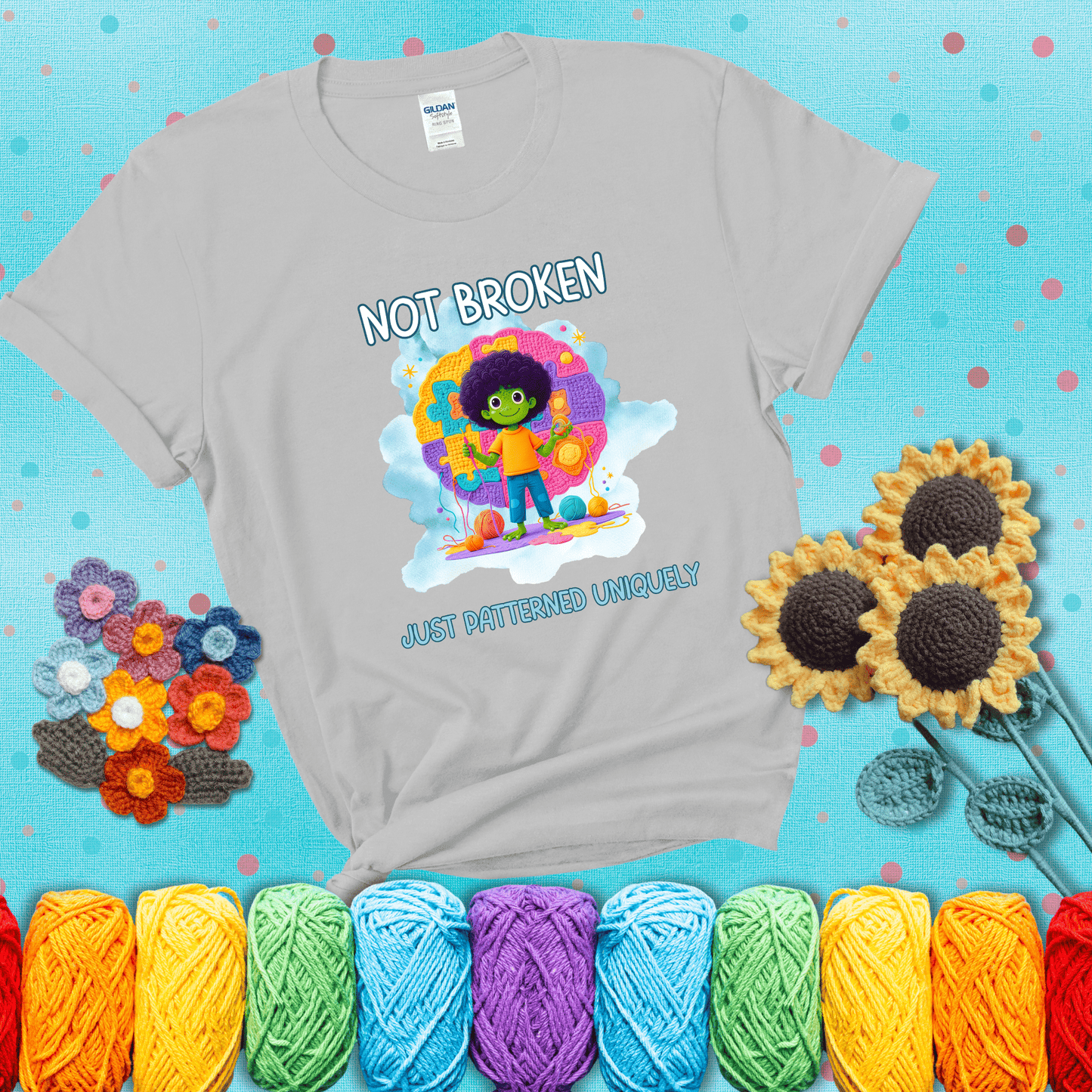 A smiling green character stands on a colorful crochet mat shaped like puzzle pieces, holding yarn and a hook, with the words “Not broken, just patterned uniquely” showing a light grey tee with a teal sunflower background