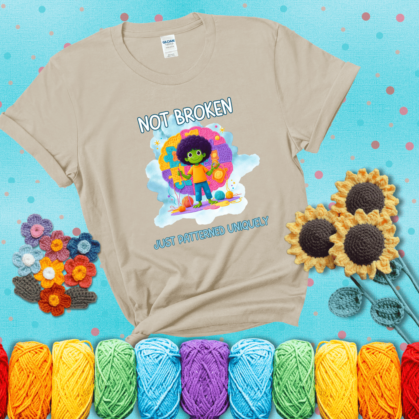 A smiling green character stands on a colorful crochet mat shaped like puzzle pieces, holding yarn and a hook, with the words “Not broken, just patterned uniquely” showing a beige tee with a teal sunflower background