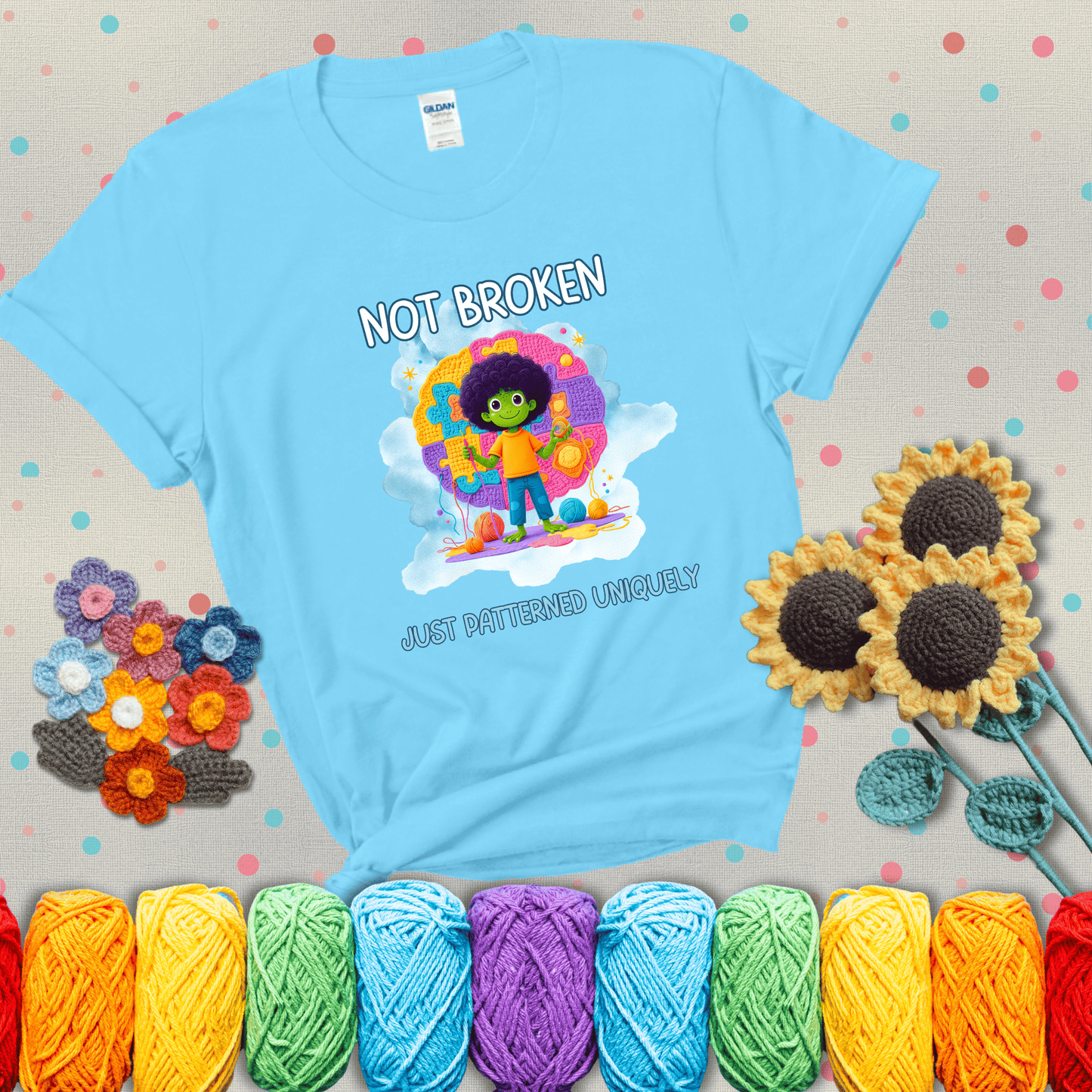 A smiling green character stands on a colorful crochet mat shaped like puzzle pieces, holding yarn and a hook, with the words “Not broken, just patterned uniquely” showing a light blue tee with grey sunflower background