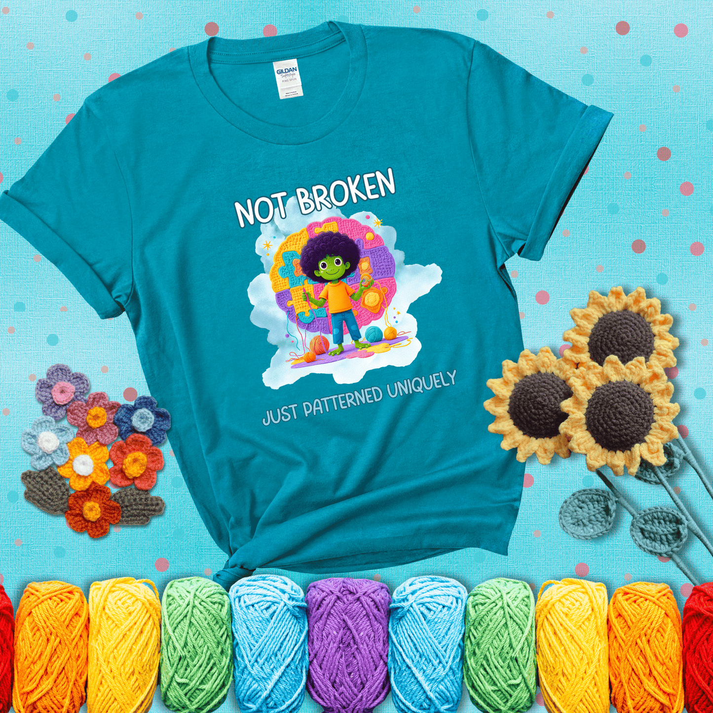 A smiling green character stands on a colorful crochet mat shaped like puzzle pieces, holding yarn and a hook, with the words “Not broken, just patterned uniquely” showing a blue-green tee with a teal sunflower background