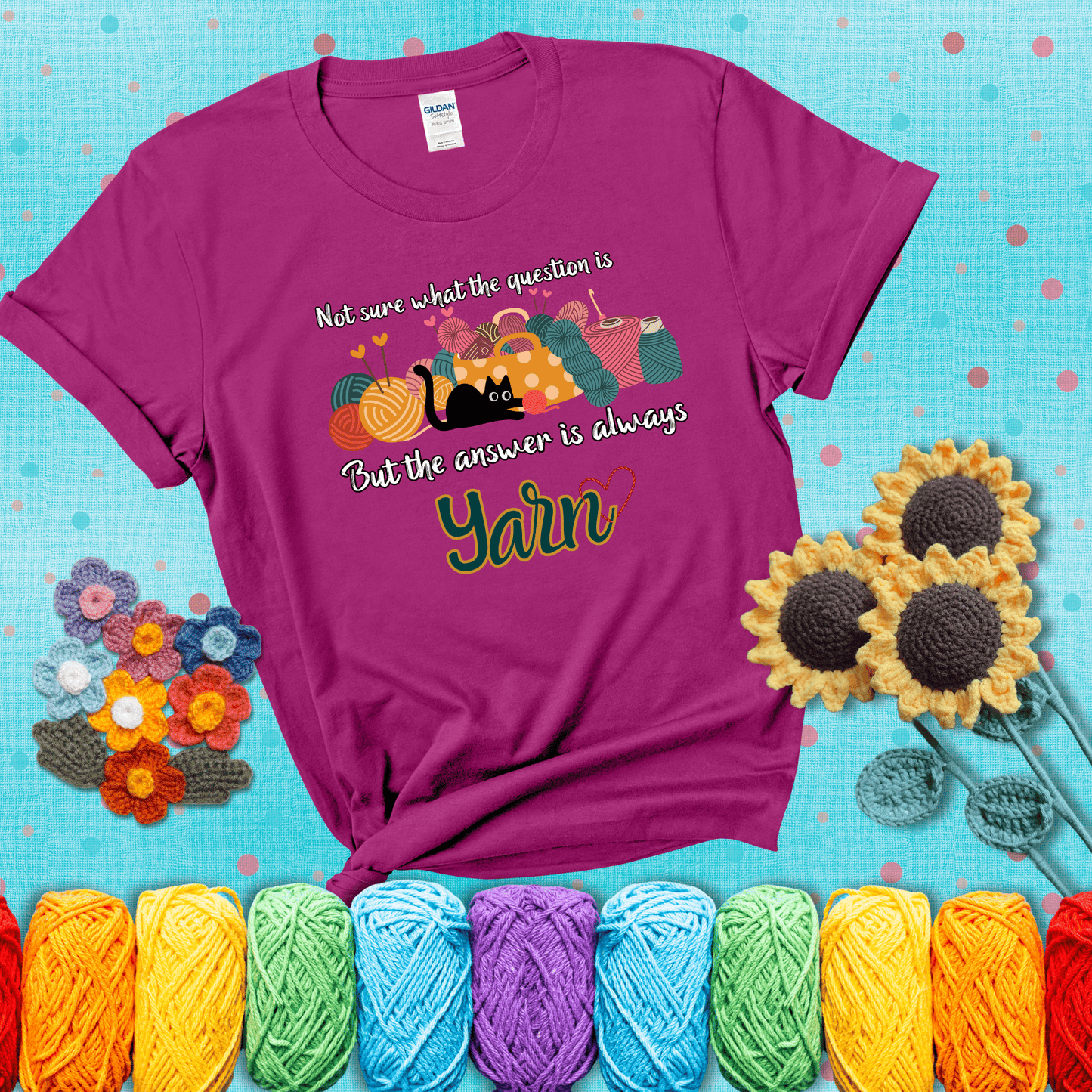 Illustration of a black cat playing with yarn next to a basket full of colorful skeins, with the phrase "Not sure what the question is, but the answer is always yarn" showing a magenta tee with a teal sunflower background