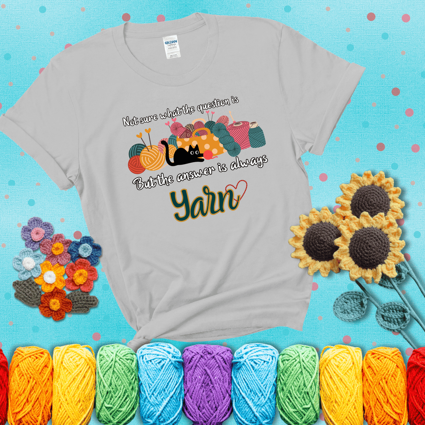 Illustration of a black cat playing with yarn next to a basket full of colorful skeins, with the phrase "Not sure what the question is, but the answer is always yarn" showing a light grey tee with a teal sunflower background