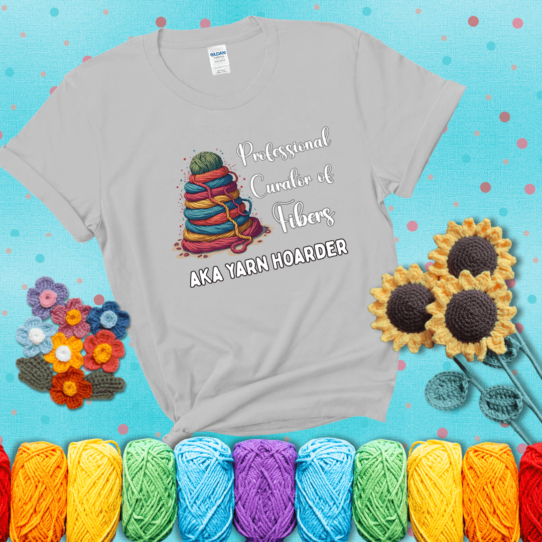 Pyramid of rainbow yarn topped with green ball; text reads “Professional Curator of Fibers AKA Yarn Hoarder,” a playful shirt for serious stashers showing a light grey tee with a teal sunflower background