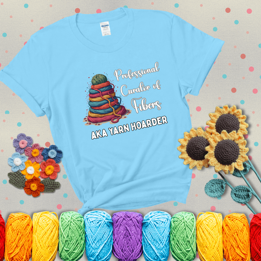 Pyramid of rainbow yarn topped with green ball; text reads “Professional Curator of Fibers AKA Yarn Hoarder,” a playful shirt for serious stashers showing a light blue tee with grey sunflower background