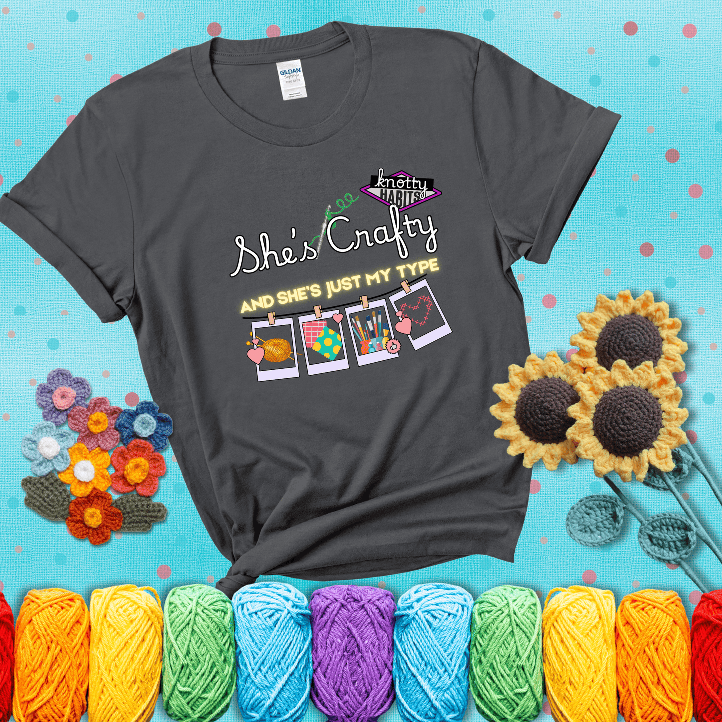 Text reads "She’s Crafty & She’s Just My Type" with yarn, fabric, painting, and cross-stitch icons hanging like Polaroids showing a dark grey tee with a teal sunflower background