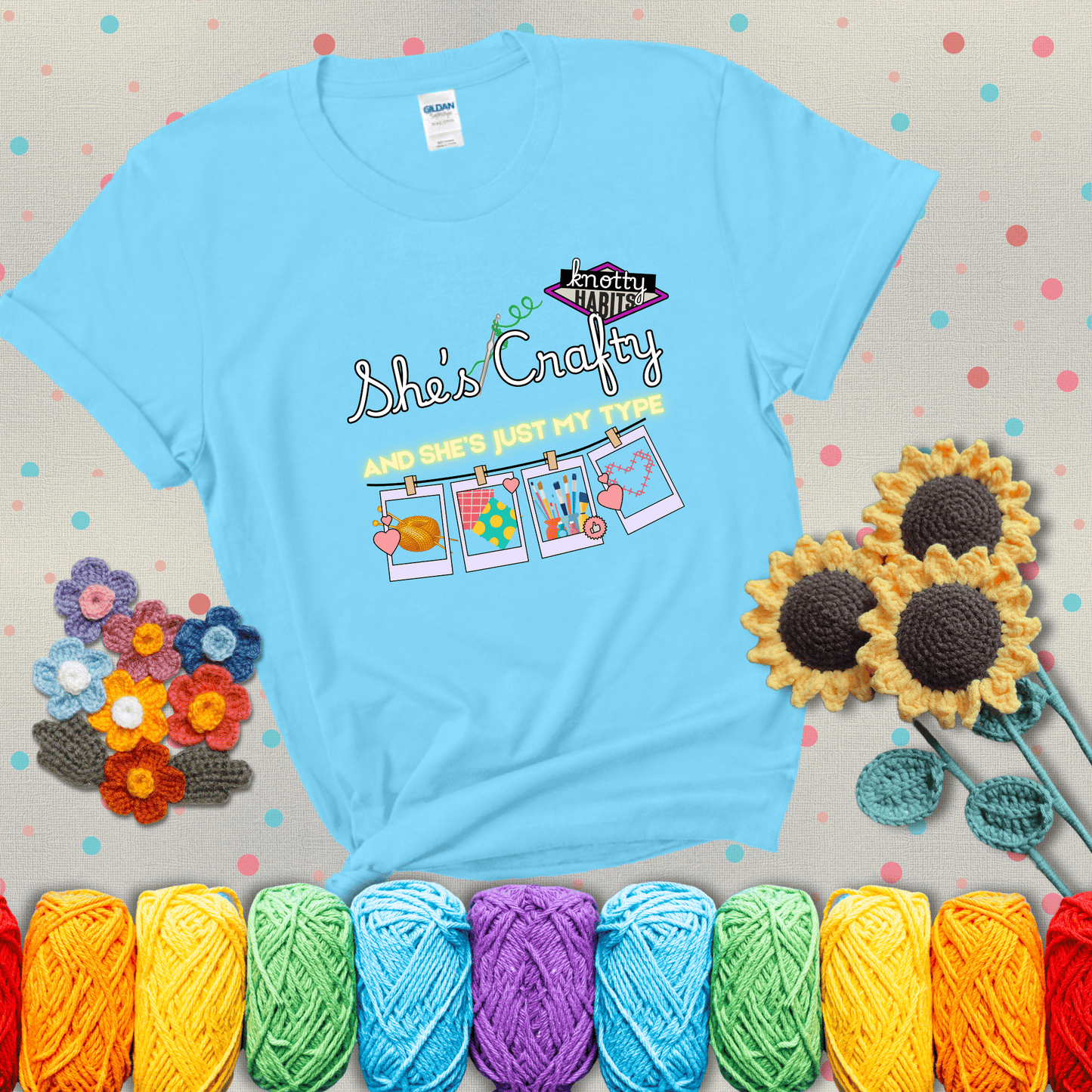 Text reads "She’s Crafty & She’s Just My Type" with yarn, fabric, painting, and cross-stitch icons hanging like Polaroids showing a light blue tee with grey sunflower background